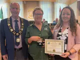 Photo of a man in a blue suit with a medal next to a woman with a green collared shirt holding an award next to a woman with curly hair and a pink blazer holding a certificate.