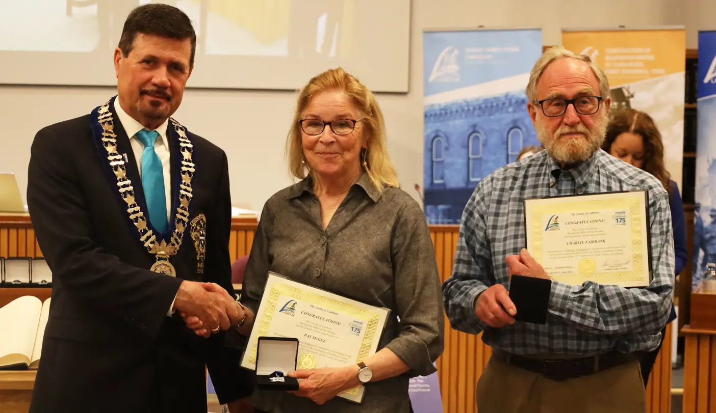 Photo of a man in a suit giving an award to a woman in a collared shirt and glasses and a man in a collared shirt and glasses.